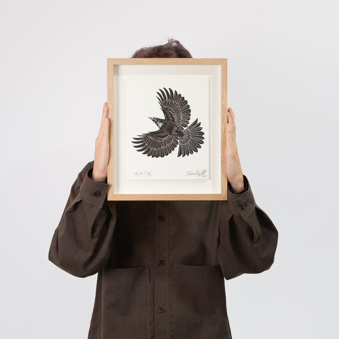 A framed black and white art print of a flying tui, featuring a light oak wood frame and a cream mat border, displayed against a neutral light gray background.