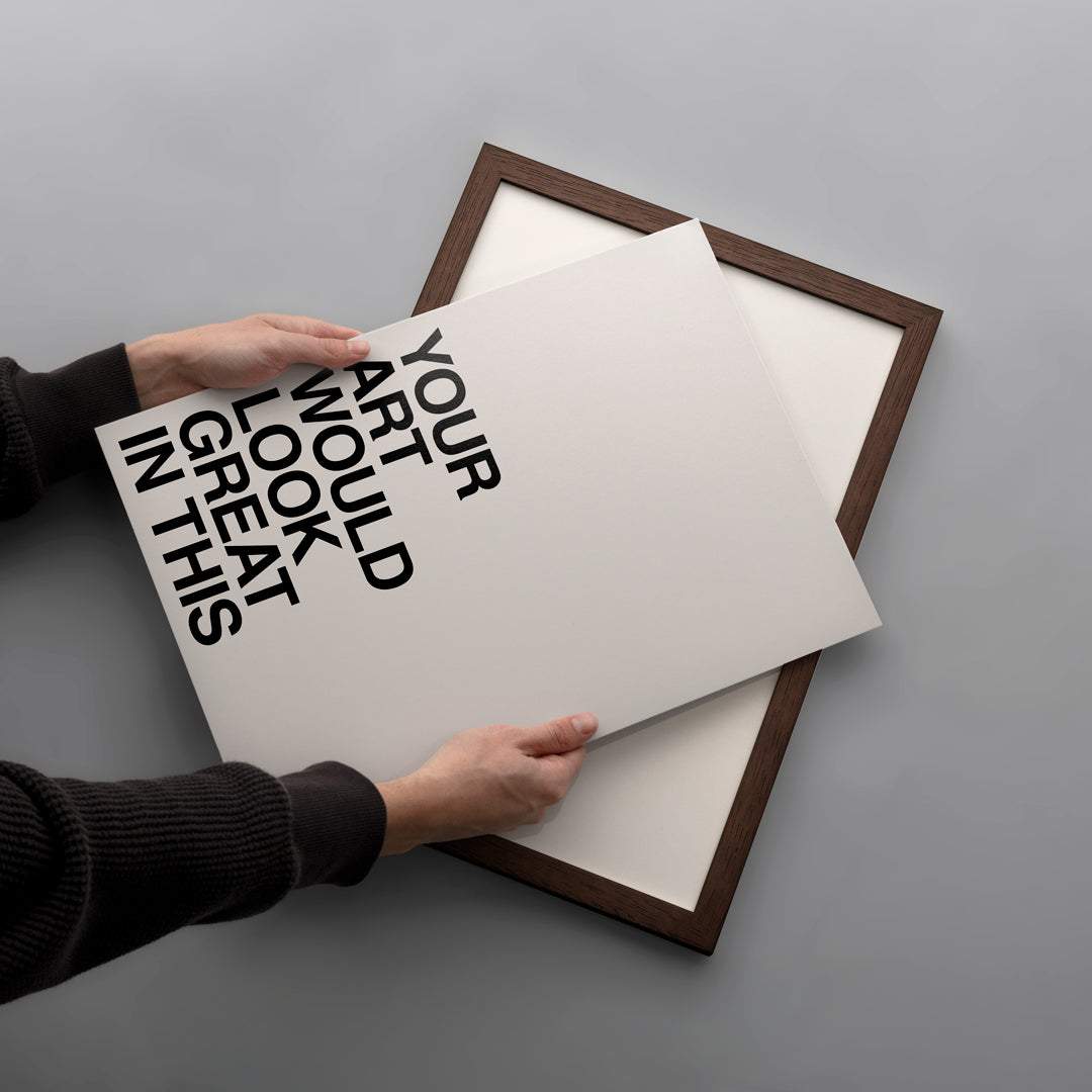 A person's hands are shown holding a white poster print with bold black typography over a dark brown wooden frame, set against a clean gray background.