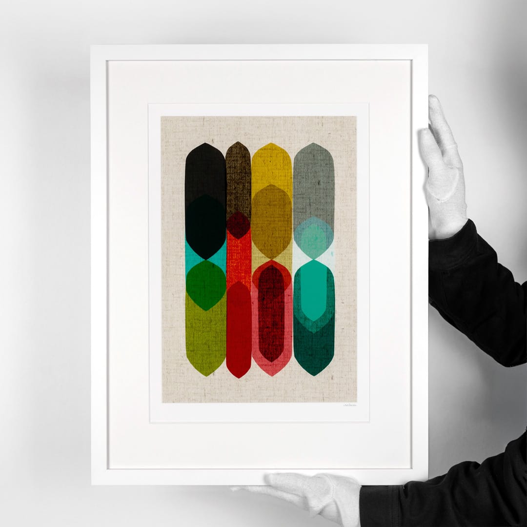 A framed abstract art print held by two hands wearing white gloves. The print features vertical, textured feather-like shapes in black, brown, yellow, red, green, and teal on a beige linen background, set within a wide white frame.