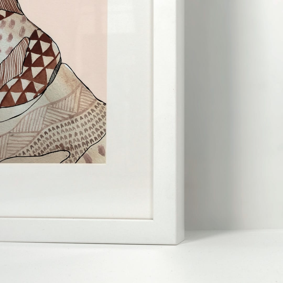 A close-up of a framed art print showing a patterned brown and beige illustration on a pale pink background, set in a clean white frame against a white wall.