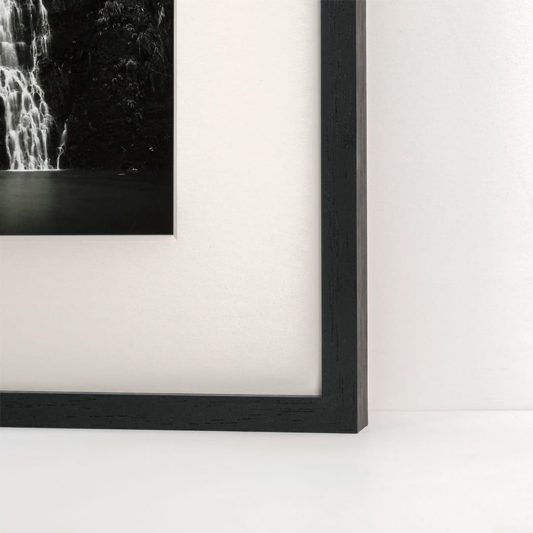 Close-up of a framed black-and-white wall art print showing part of a waterfall scene, with a thick black frame against a white wall and floor.